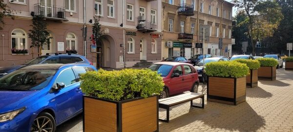 Ławka i duże, drewniane donice z roślinnością, stojące na chodniku.