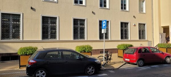 Duże, drewniane donice z roślinnością, stojące obok miejsc parkingowych.