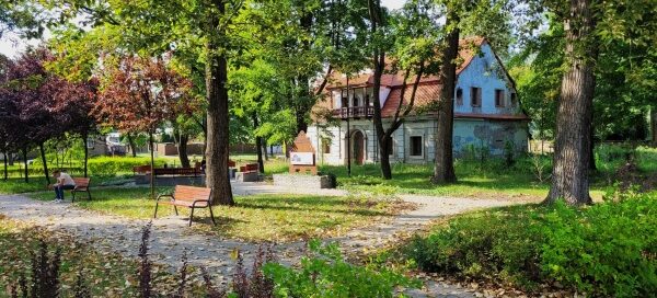 Ścieżki i alejki wśród drzew. W tle widoczny dworek.