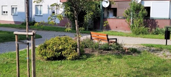 Zielony skwer przy którym stoi ławka. W tle widać blok.