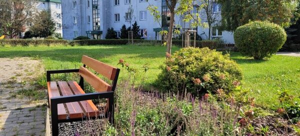 Zielony skwer przy którym stoi ławka.