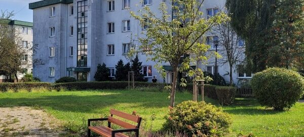 Zielony skwer przy którym stoi ławka. W tle widać blok.