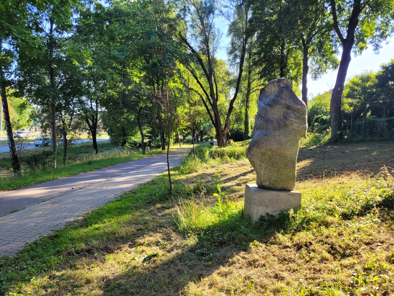 Chodnik i ścieżka rowerowa, biegnące wśród wiekowych drzew. Na trawniku ustawione Rzeźby.