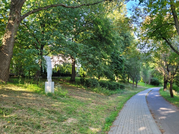 Chodnik i ścieżka rowerowa, biegnące wśród wiekowych drzew. Na trawniku ustawione Rzeźby.