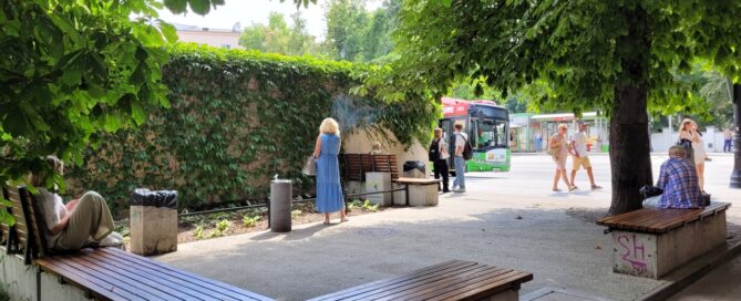 Betonowy skwer z ławkami, otoczony roślinnością.