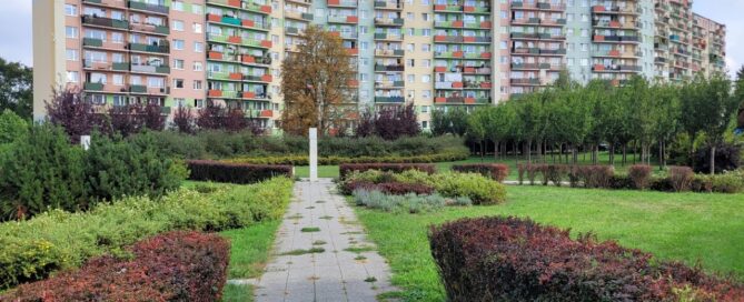 Skwer z roślinnością. W tle widoczny blok mieszkalny.