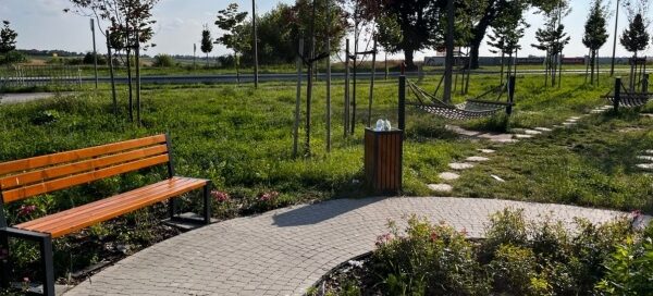 Brukowana alejka skweru, przy której ustawione są kosz i ławka.