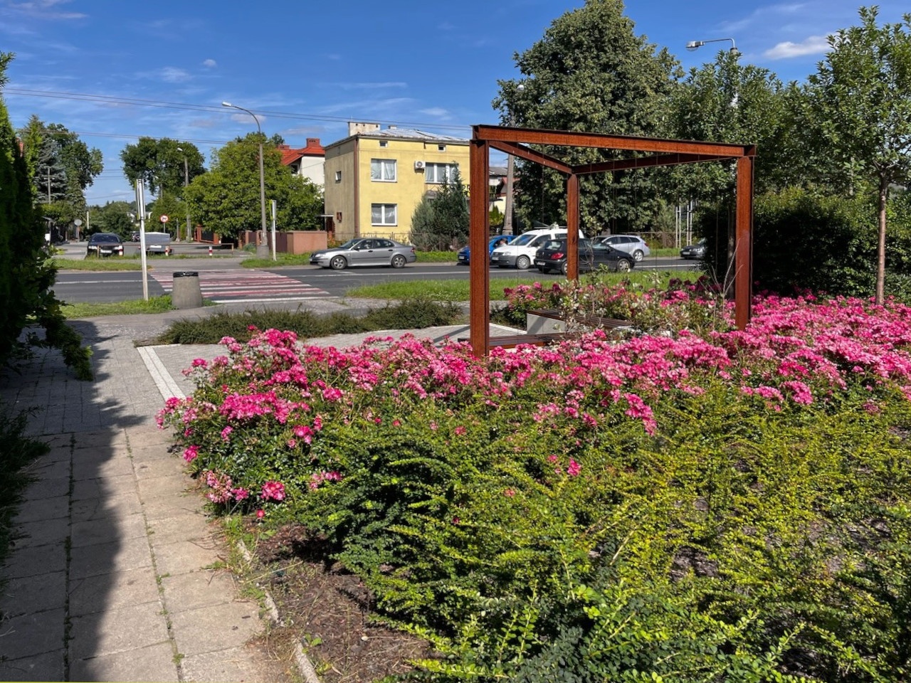 Mały skwer z betonowymi ławkami i pergolą, ozdobioną roślinnością.