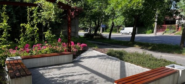 Mały skwer z betonowymi ławkami i pergolą, ozdobioną roślinnością.