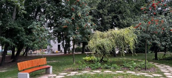 Ścieżka z nieregularnych płyt w trawniku, otoczona starodrzewami. Obok ścieżki postawiono ławkę.