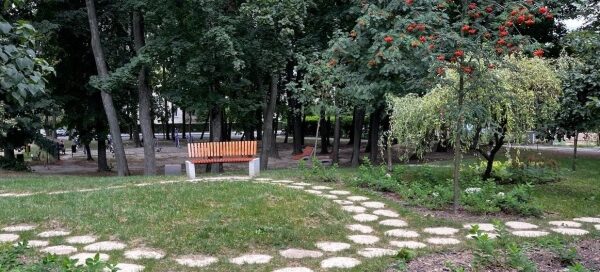 Ścieżka z nieregularnych płyt w trawniku, otoczona starodrzewami. Obok ścieżki postawiono ławkę.