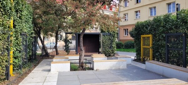 Betonowe ławki i metalowe płotki, otoczone żywopłotem.