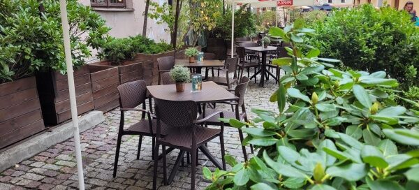 Stoliki na zewnątrz restauracji otoczone roślinnością w donicach.