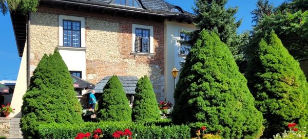Brukowane alejki, drzewa, kwitay i żywopłot. W tle widoczny stary dwór.
