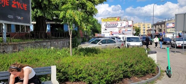 Mały skwer z nasadzeniami roślinnymi, ławką i koszem.