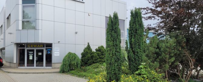 Wydzielony teren przed wejściem do siedziby firmy, ozdobiony nasadzeniami krzewów i drzew.