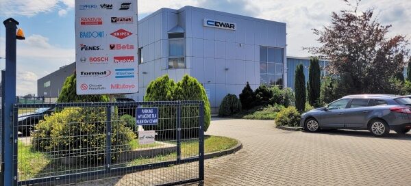 Brama wjazdowa na teren firmy. Przy wjeździe widoczne są krzewy i drzewa.
