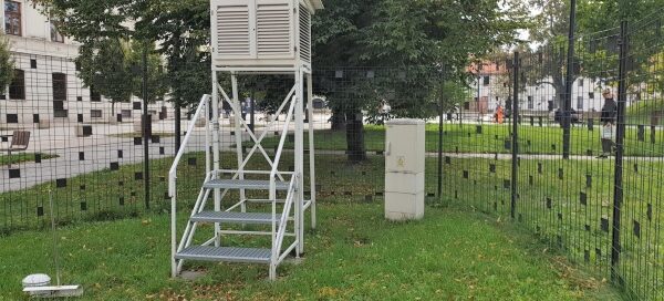 Stacja meteorologiczna. Przęsła ogrodzenia ozdobione są czarnymi kwadratami, które pełnią rolę maskującą. W tle widoczne wysokie drzewa.