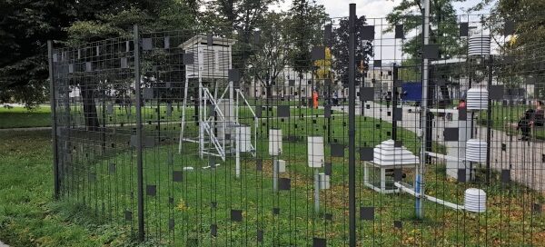 Stacja meteorologiczna. Przęsła ogrodzenia ozdobione są czarnymi kwadratami, które pełnią rolę maskującą. W tle widoczne wysokie drzewa.