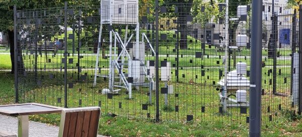 Stacja meteorologiczna. Przęsła ogrodzenia ozdobione są czarnymi kwadratami, które pełnią rolę maskującą. W tle widoczne wysokie drzewa.