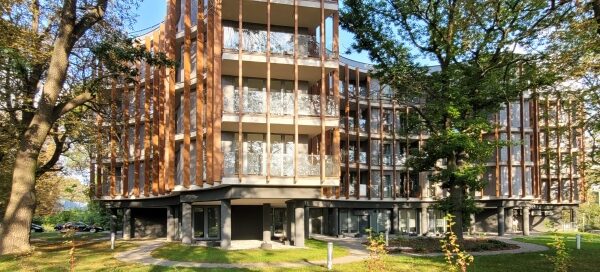 Nowoczesny budynek mieszkalny, wkomponowany między starymi drzewami. Widoczne ścieżki dla pieszych, zaprojektowane na terenie trawnika.