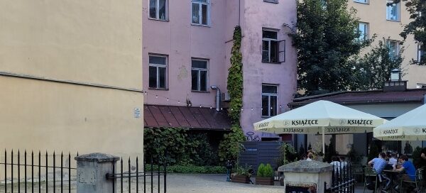 Podwórze kamienicy, gdzie mieści się lokal gastronomiczny. Miejsce ze stolikami otoczone jest roślinnością w donicach, a w rogu budynku widoczne są pnącza.