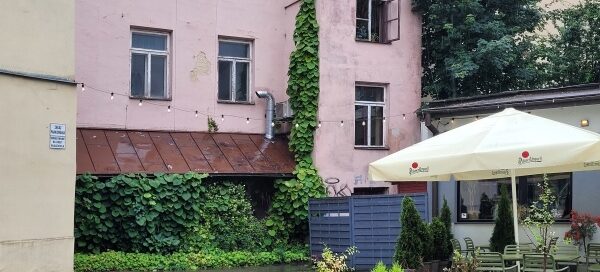 Podwórze kamienicy, gdzie mieści się lokal gastronomiczny. Miejsce ze stolikami otoczone jest roślinnością w donicach, a w rogu budynku posadzono pnącza.