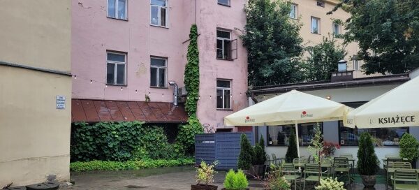 Podwórze kamienicy, gdzie mieści się lokal gastronomiczny. Miejsce ze stolikami otoczone jest roślinnością w donicach, a w rogu budynku widoczne są pnącza.