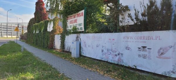 Płot z banerami reklamowymi porośnięty pnączami.