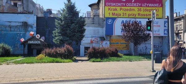 Teren trawnika z nasadzeniami krzewów i drzew. W tle widoczny budynek i banery reklamowe.