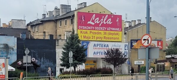 Teren trawnika z nasadzeniami krzewów i drzew. W tle widoczny budynek i banery reklamowe.