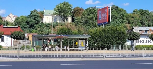 Jezdnia i przystanek autobusowy na tle wzgórza porośniętego drzewami.