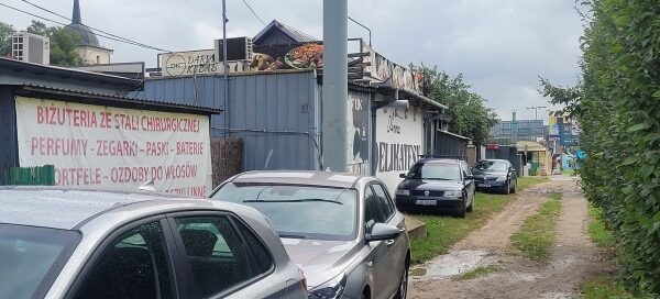 Samochody zaparkowane na terenie trawnika za budami usługowymi.