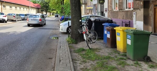Wydzielone trawniki oraz drzewa rosnące przy chodniku. Widoczne kolorowe pojemniki na śmieci.