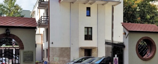 Miejsce z parkingiem i białym, modernistycznym budynkiem w głębi. Balkon z pnączami mieści się na ostatnim piętrze budynku.