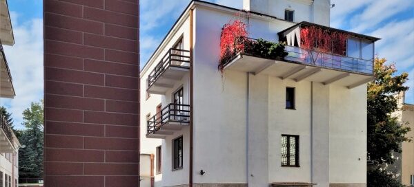 Biały budynek, na którego, ostatnim piętrze, mieści się balkon z pnączami.