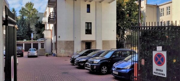 Miejsce z parkingiem i białym, modernistycznym budynkiem w głębi. Balkon z pnączami mieści się na ostatnim piętrze budynku.