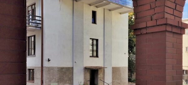 Biały budynek, na którego, ostatnim piętrze, mieści się balkon z pnączami.