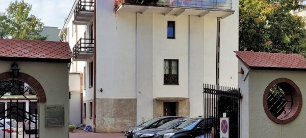 Miejsce z parkingiem i białym, modernistycznym budynkiem w głębi. Balkon z pnączami mieści się na ostatnim piętrze budynku.