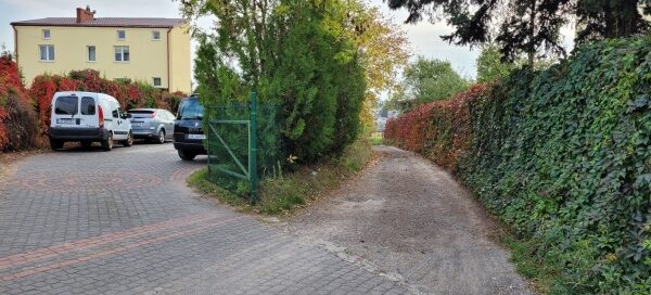 Prywatna posesja z budynkiem mieszkalnym. Obok niej widoczna droga gruntowa po obu stronach otoczona ogrodzeniem, na którym zasadzono pnącza.