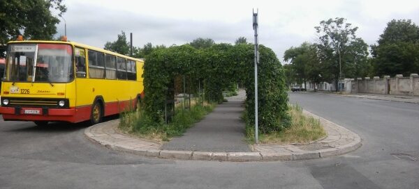 Pergole ustawione nad chodnikiem dla pieszych, obok przystanku autobusowego. Tworzą bramę z roślin nad chodnikiem.