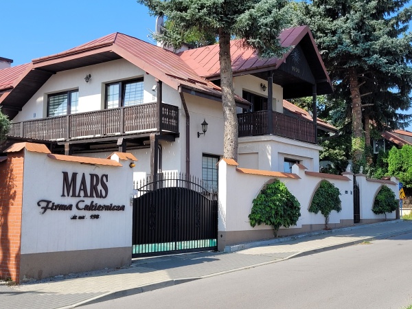 Budynek z dwiema starymi sosnami, otoczony murem, w którego otworach zasadzono pnącza. Pnącza mają formę drzewek.