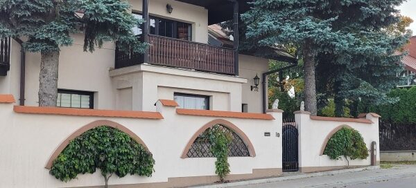 Budynek i mur, w którego otworach zasadzono pnącza. Pnącza mają formę drzewek.