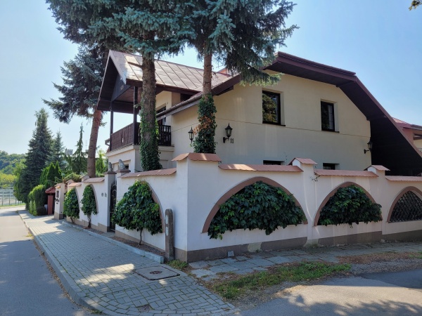 Budynek z trzema starymi sosnami, otoczony murem, w którego otworach zasadzono pnącza. Pnącza mają formę drzewek.