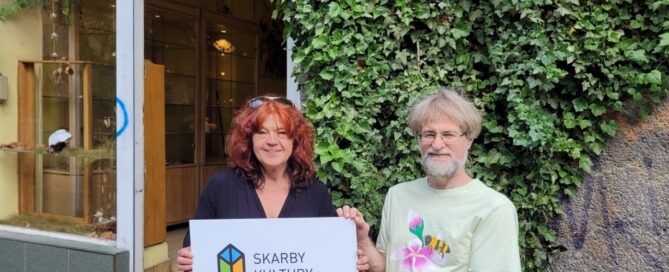 Dwie osoby trzymające tabliczkę z logo Skarby Kultury Przestrzeni, na tle lokalu, którego wejście ozdobione jest pnączami.