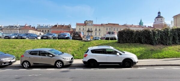 Zarośnięty pnączami narożnik parkingu przy skrzyżowaniu ulic. W tle widoczne kamienice.