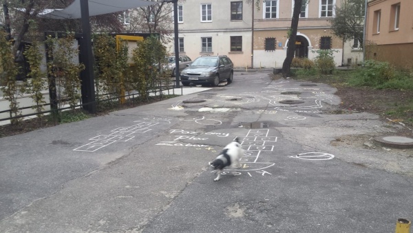 Miejsce, gdzie jest namalowany poziomy plac zabaw. Asfaltowa droga. W oddali widoczne zabudowania miejskie.