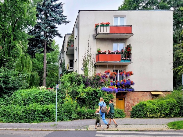 Blok mieszkalny usytuowany wśród zieleni. Widoczne drzewa i krzewy oraz balkony ozdobione kwiatami.