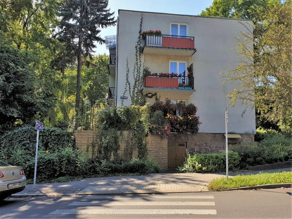 Blok mieszkalny usytuowany wśród zieleni. Widoczne drzewa i krzewy oraz balkony ozdobione kwiatami.